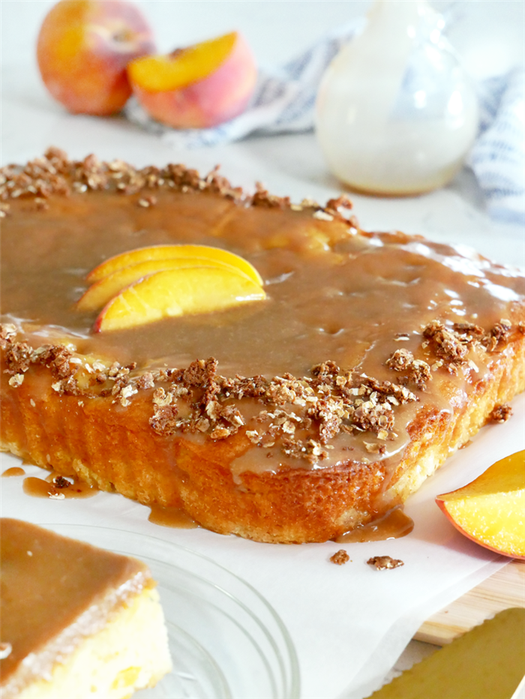 Peach sheet cake topped with a brown sugar caramel glaze and slices peaches with pecans and oat crumbled on top.