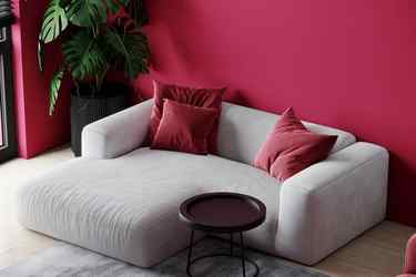 A section of a living room with a comfortable rounded couch and a coffee table. The pillow and walls are magenta, reflecting Pantone’s Color of the Year 2023: Viva Magenta.