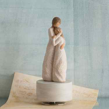 Two faceless girl figurines in white dresses hugging one another - standing on round white plaque with blue background and music sheet below them