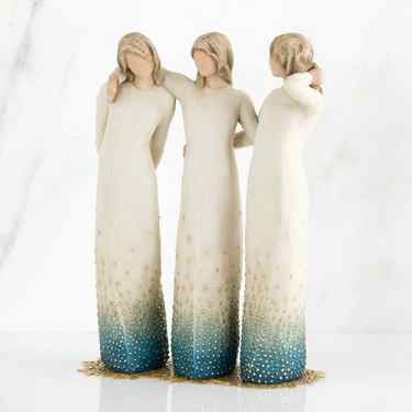 Set of three standing faceless woman figurines wearing white dresses with blue on the ends of them - they all have their arms around each other