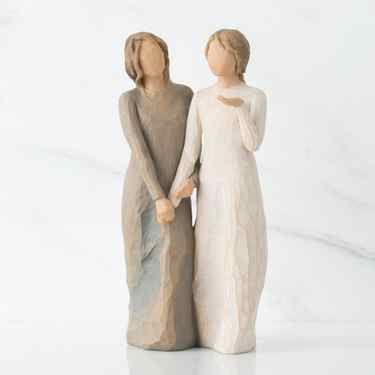 Two faceless woman figurines standing facing forward holding hands - left one is wearing light brown dress, right one is wearing white dress