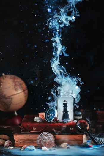 A still life photography arrangement of a lighthouse silhouette in a smoking glass jar with books, a compass, magnifying glass, and globe against a dark background