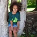 Load image into Gallery viewer, Female wearing dark green Unisex Youth Camp Scene T-shirt. Bright green Camp Scene logo screened on centre chest. 