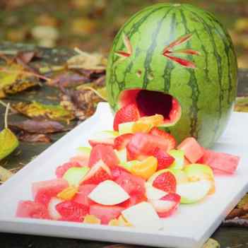 A white tray on a leaf covered picnic table containing a jack-o-lantern watermelon with fruit salad spewing out of its mouth.