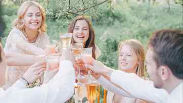 Dinner party guests toasting with their drinks, wearing white and sitting outside