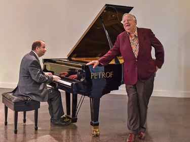 Seward Johnson rests his hand on an open grand piano singing while a pianist plays